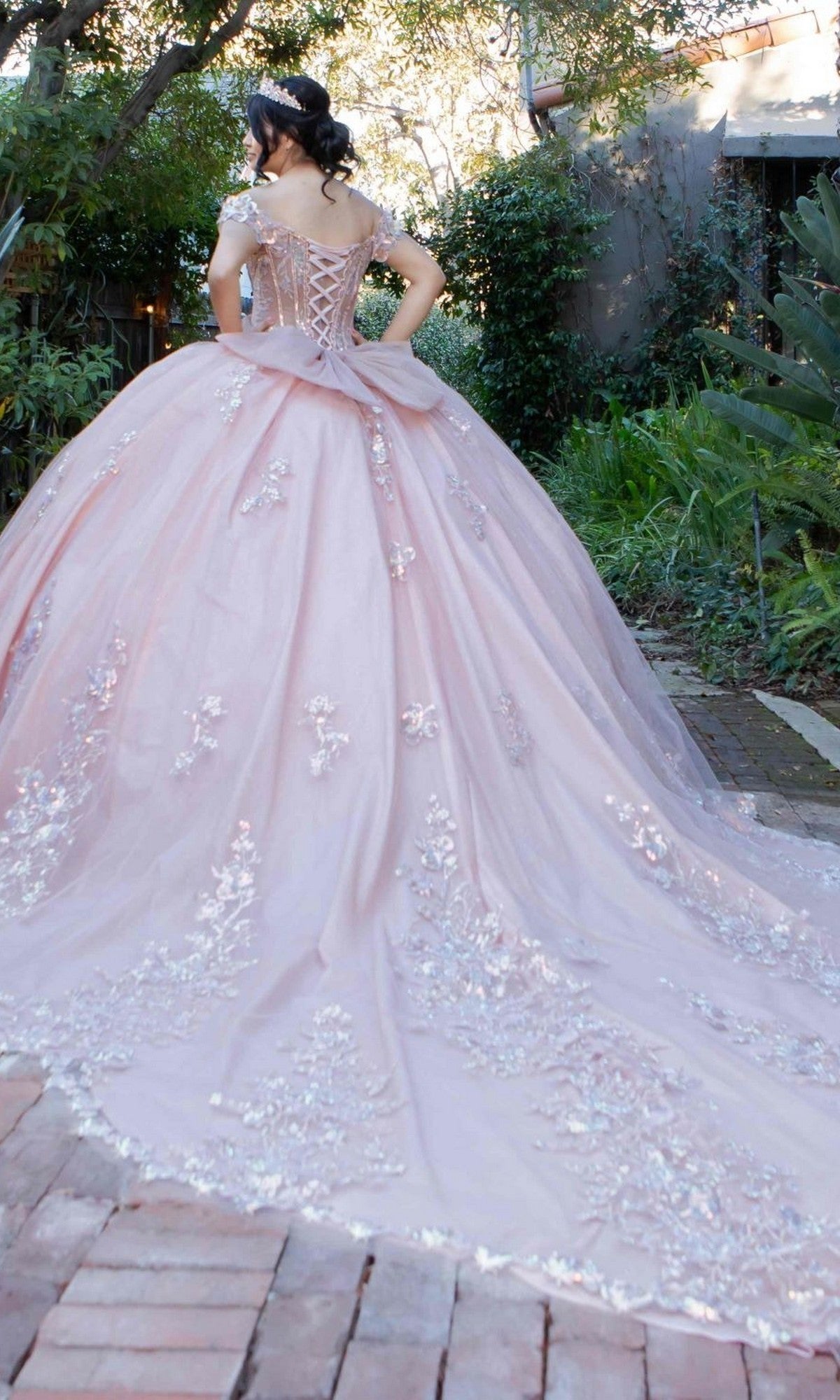 A woman stands outdoors facing away from the camera, wearing the Quinceanera Dress 8370J By Cinderella Couture—a light pink ball gown adorned with floral embroidery and delicate floral appliques, featuring an illusion corset bodice and a long train.